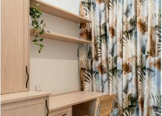 A minimalist home office setup featuring light wood cabinetry, a built-in desk, a rattan chair with chrome legs, and leafy patterned floor-to-ceiling curtains. Floating shelves hold a trailing plant, adding a touch of greenery to the neutral space.