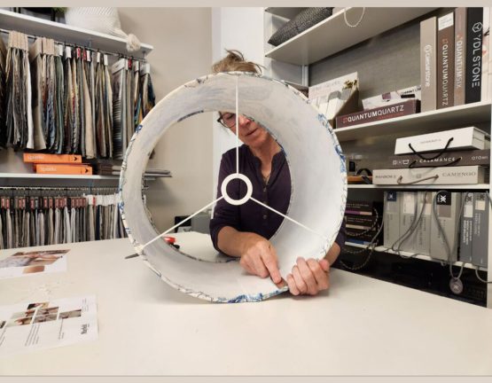 Person assembling a lampshade in an interior design workshop with fabric samples in the background.